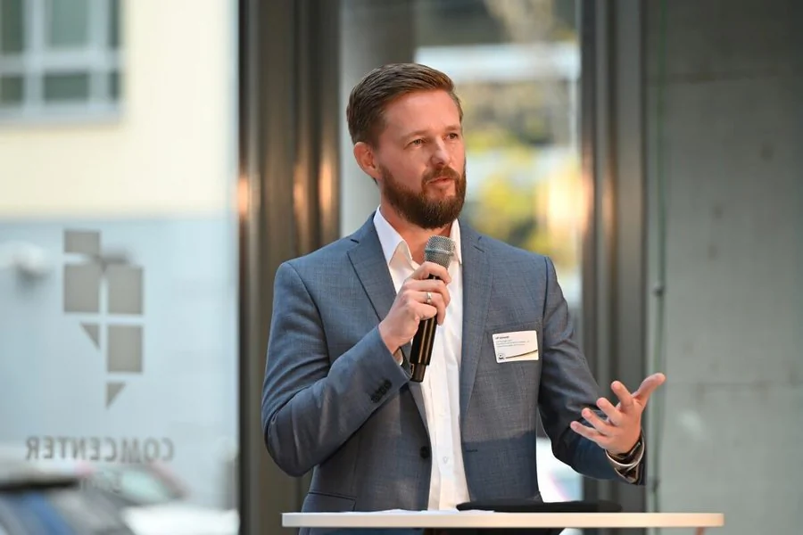 Ein bärtiger Mann in einem Anzug hält ein Mikrofon und spricht zu einem Publikum.