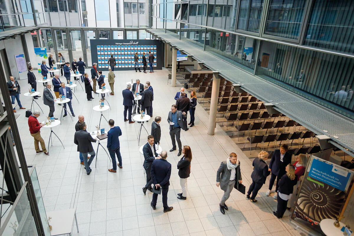 Bildbeschreibung / description: Eine Ansammlung von Menschen steht in einem geräumigen Gebäude und führt angeregte Gespräche.