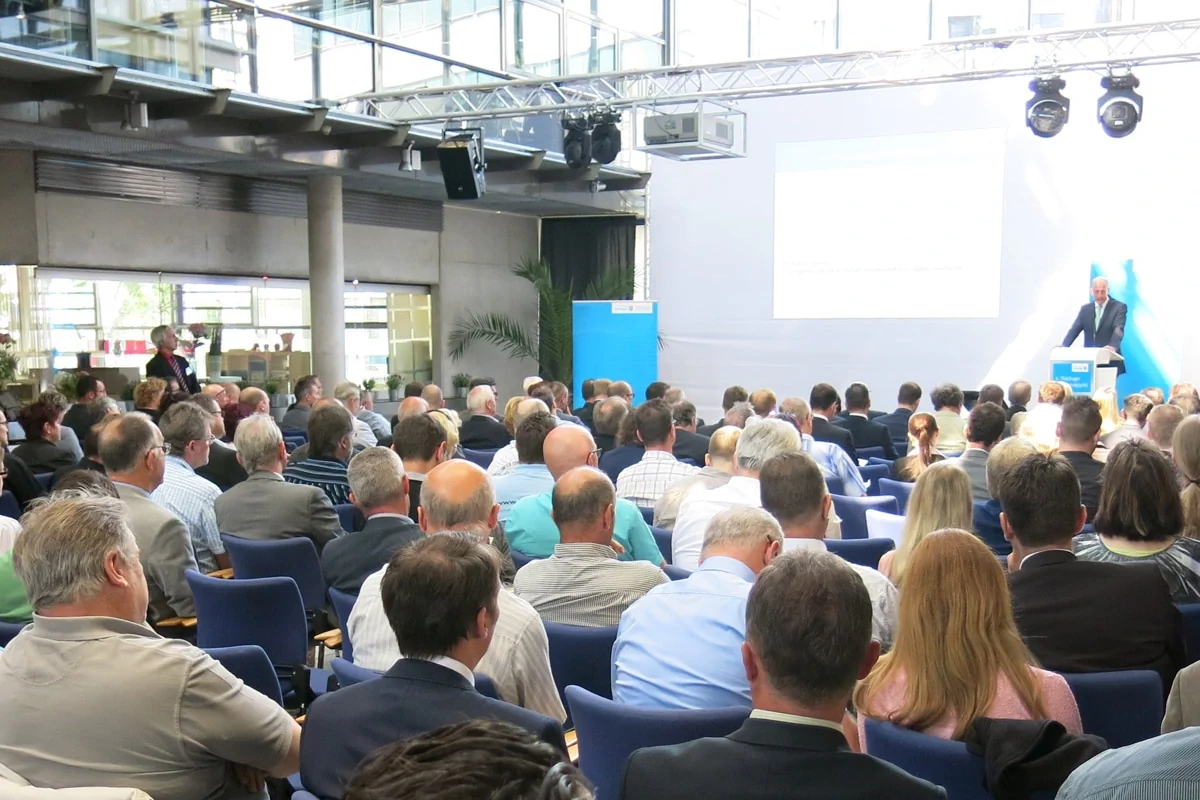 Bildbeschreibung / description: Eine Vielzahl von Menschen ist in einem großen Raum auf Stühlen versammelt und lauscht einem Vortrag